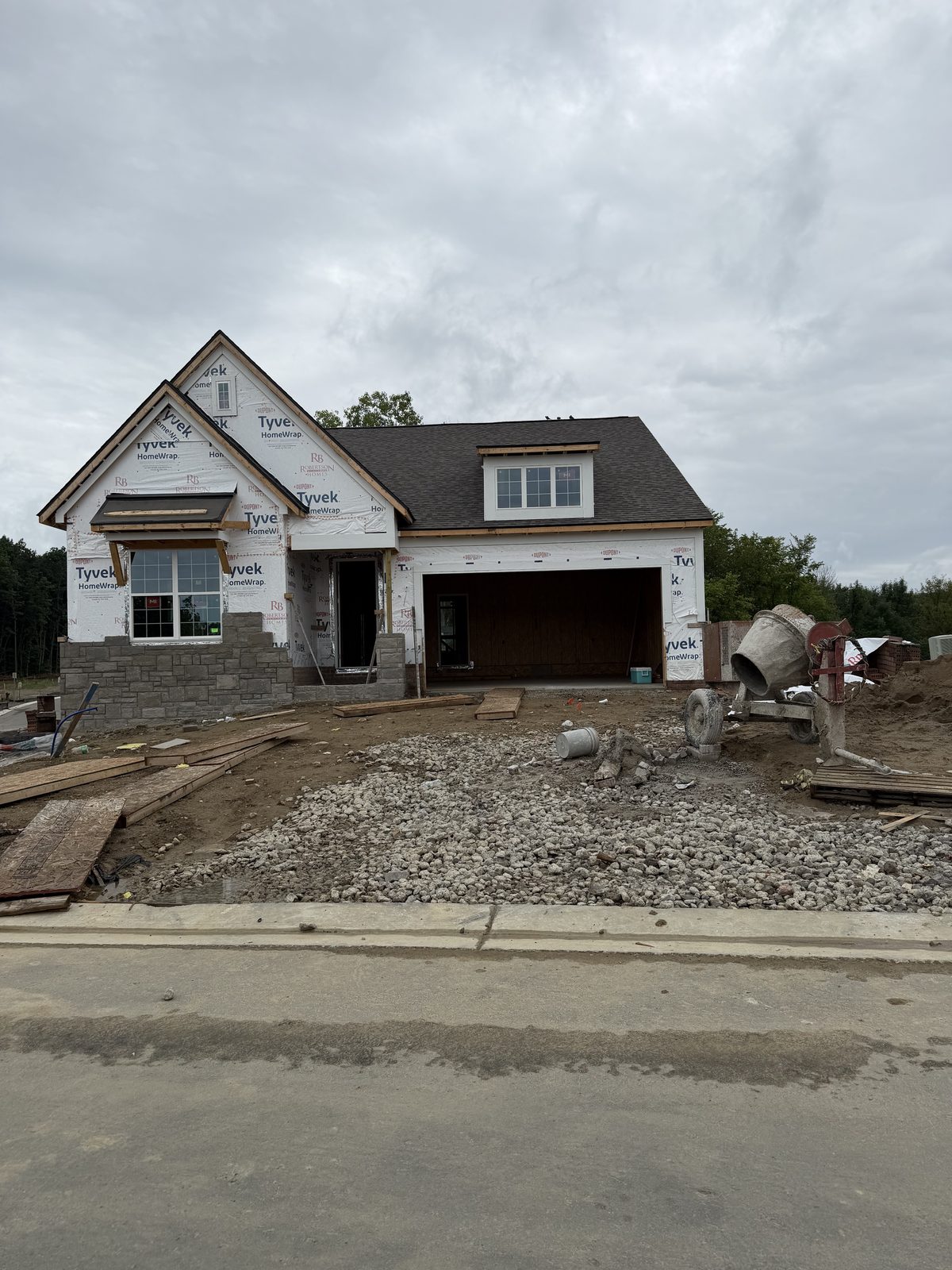 A new-construction home under construction in the South Lyon area, with house wrap, a shingled roof, and partial stone veneer on a gravel lot.