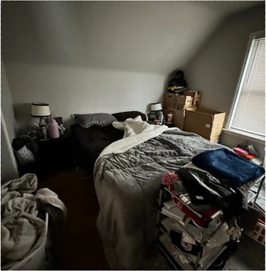 Primary bedroom before staging: unmade grey bedding, moving boxes stacked by the window, a clothing rack at the foot of the bed, and everyday clutter throughout the room