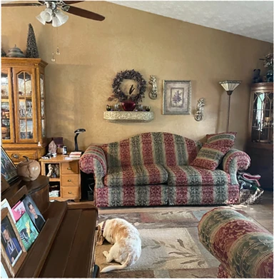 Traditional living room before staging: busy patterned sofa, oak china cabinet, wall décor, floor lamp, and personal items throughout the seating area