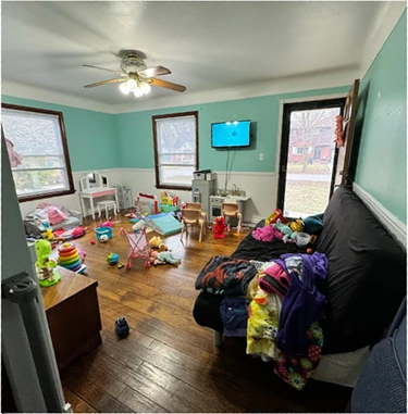 Living room before staging: cluttered playroom with toys scattered across hardwood floors, teal upper walls, white wainscoting, and a sofa covered with blankets and clothing